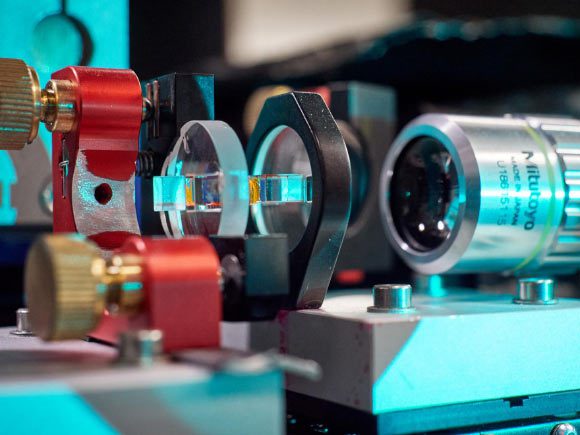 Le microrésonateur optique rempli d'une solution de colorant (jaune) ; à droite, un objectif de microscope utilisé pour observer et analyser la lumière émergeant du résonateur. Crédit image : Gregor Hübl / Université de Bonn.