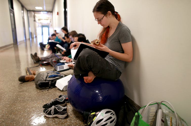 Une étudiante est assise sur une balle de yoga dans un couloir et lit un livre. D'autres étudiants sont sur des ordinateurs portables derrière elle.