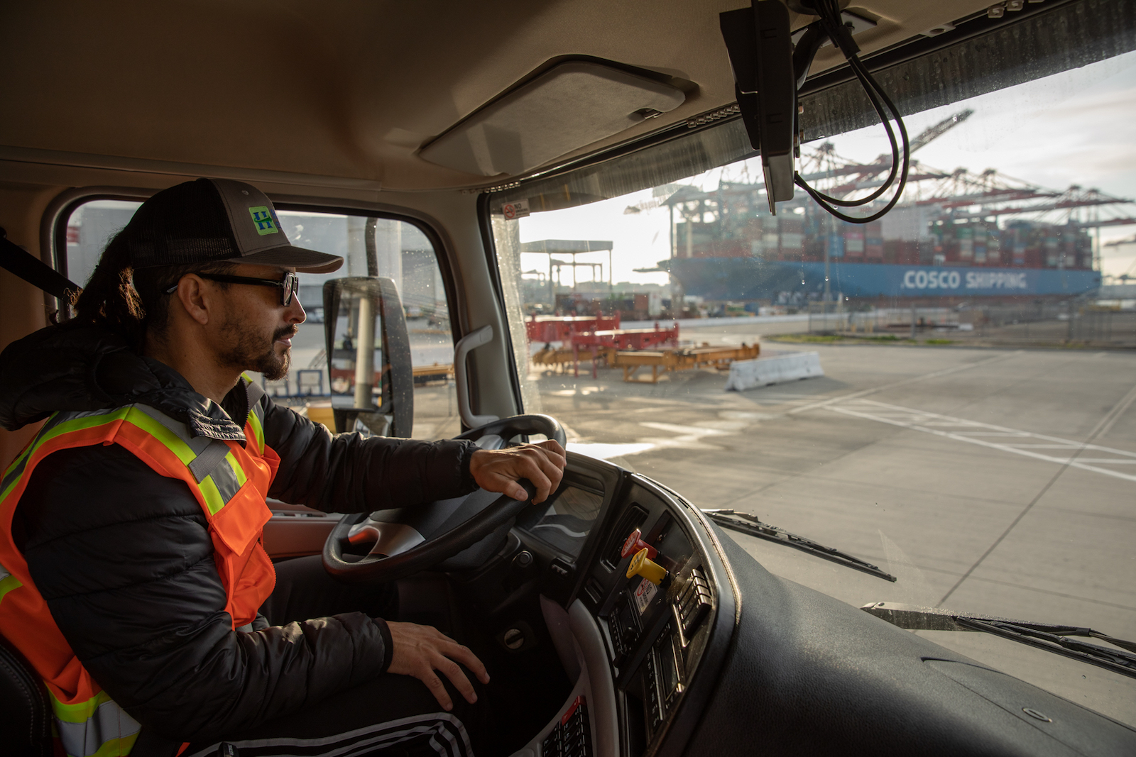 un homme vêtu d'un gilet orange et d'un chapeau de camionneur est assis à l'intérieur de la cabine d'un gros camion roulant près de conteneurs d'expédition