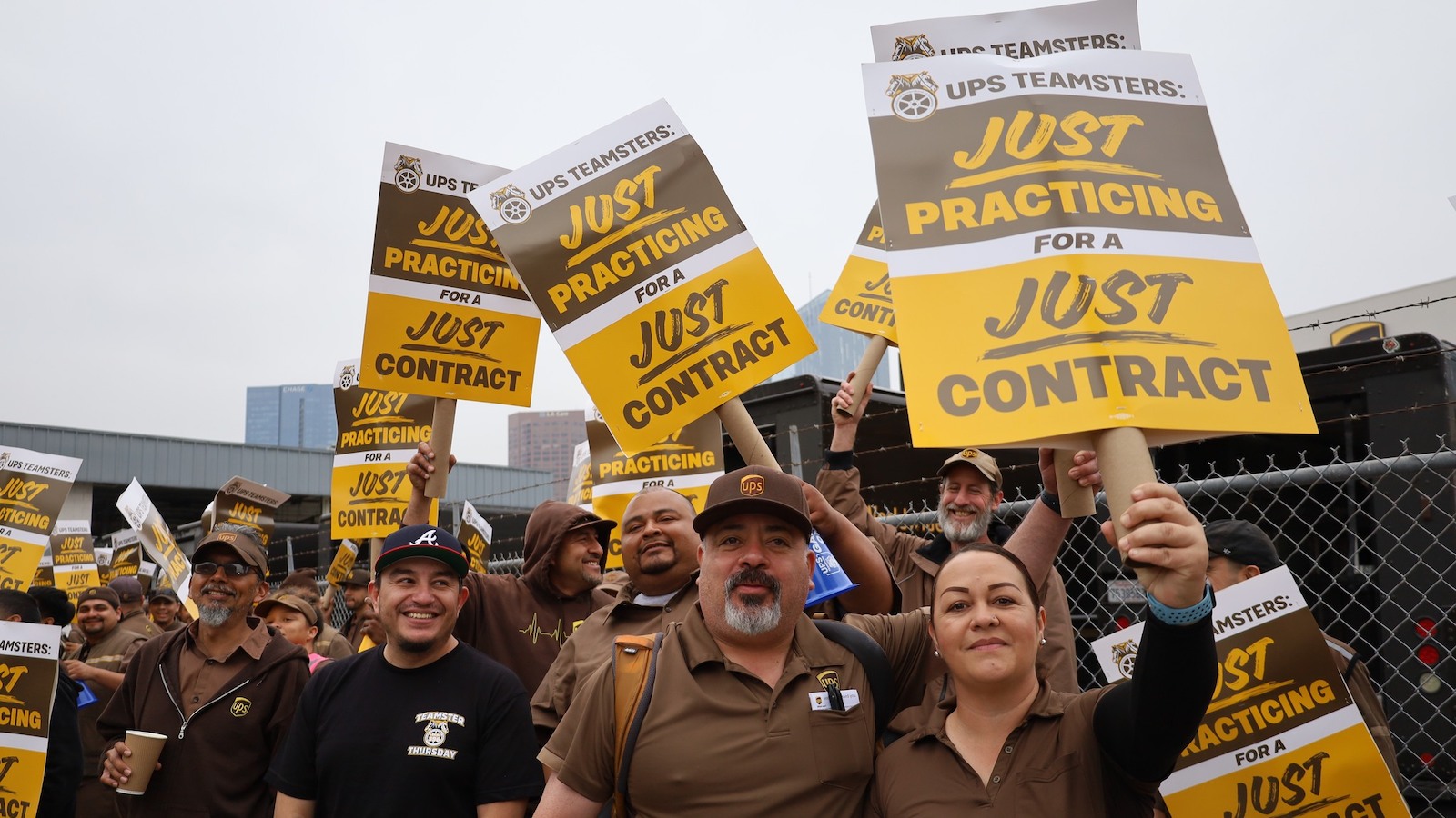 Travailleurs d'UPS en uniformes marrons debout à l'extérieur d'un entrepôt, tenant des pancartes indiquant 